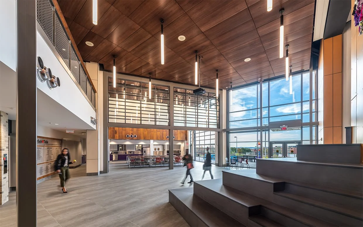 Wood ceiling and wall paneling above a café beside tiered seating in a double-height interior.
